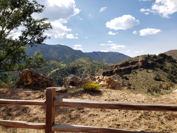 A view of the mountains from Cave of the Winds, Manitou Springs. Small fluffy clouds dot the sky. Rusty red sandstone peaks out from under the brush. The very distant chaparral looks blue in the thin haze blown from the west wildfires.