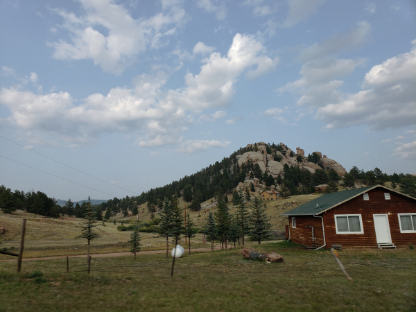 Outside of Colorado Springs, the town of Divide, Colorado, 45 minutes northwest. A small cabin sits right at the foot of mountain that looks more like a heap of boulders, littered with trees. Another small, red wooden house sits in the foreground. It has white doors and windowframes and looks shabby, but proud.