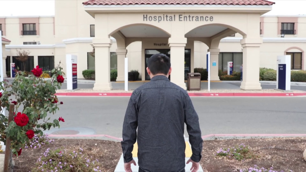 A lone man in a dark shirt who definitely seems like he has problems stands in front of a hospital entrance.