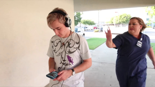 A nurse runs up to a young man attempting to enter the building, unable to hear her as he is listening to loud music on his headphones connected to his phone.