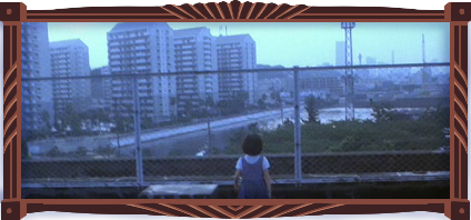 Little Ikuko in her blue overalls stands behind the chainlink fence of the apartment rooftop. The sky is bright: But not clear, and it’s eerie. You can see the river from here, dammed by concrete.